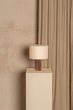 Table lamp with wooden base and beige shade on a white pedestal against a beige wall and curtain.