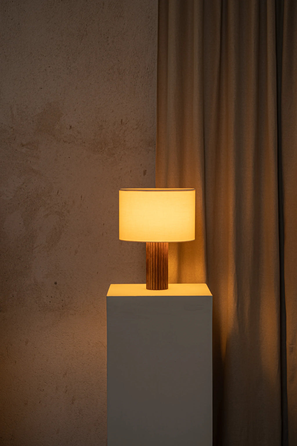 Table lamp with wooden base on a gray pedestal against a textured wall and curtain.