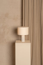 Beige lamp on a white pedestal against a beige wall with curtains.