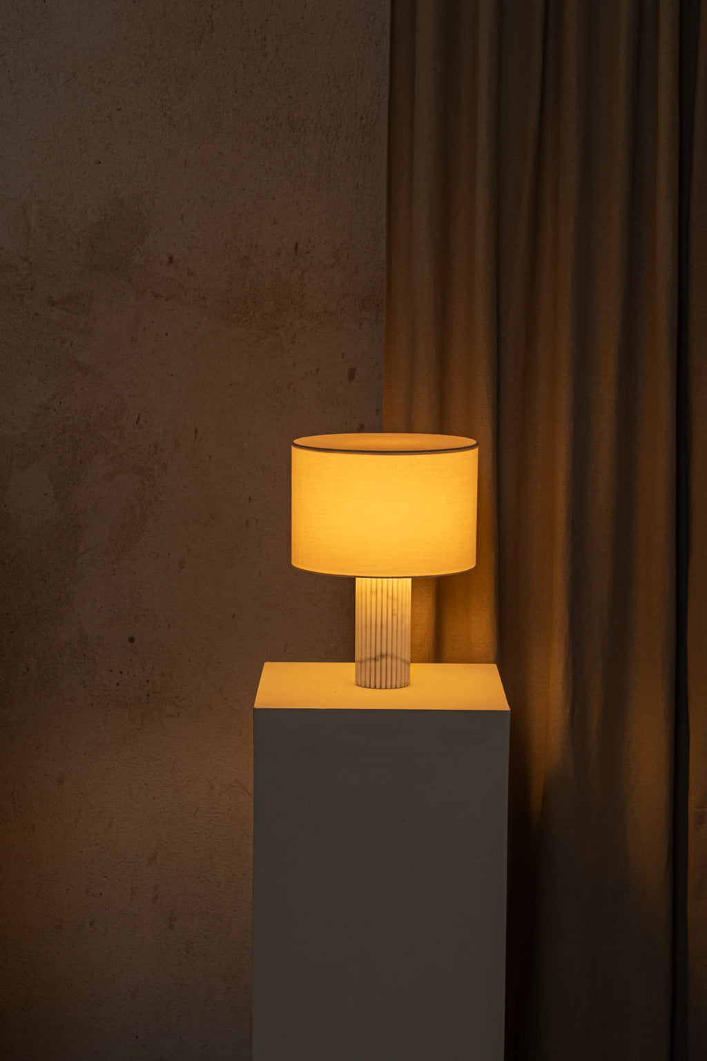 Table lamp with a warm glow on a gray pedestal against a textured brown wall.