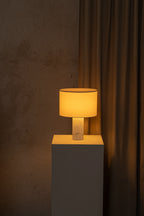 Table lamp with a warm glow on a gray pedestal against a textured brown wall.