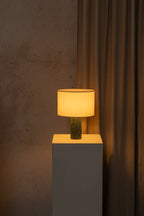 Table lamp with a textured shade on a gray pedestal against a brown curtain.