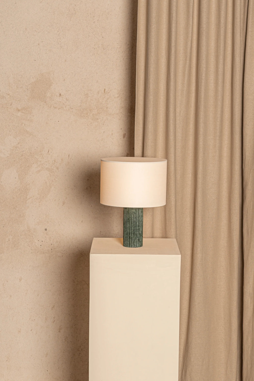 Table lamp with a green base on a white pedestal against a beige wall and curtain.