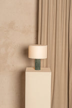 Table lamp with a green base on a white pedestal against a beige wall and curtain.