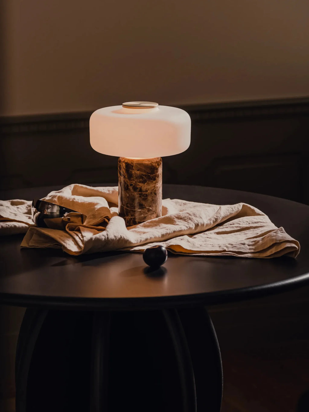 TAMO Fonte Table Lamp: This modern luxury lamp features a cylindrical brown marble base with white veining, topped by an illuminated luminous white glass shade and a small brass cap. Displayed on a dark table with a light draped linen cloth, sunglasses, and a black stone. Ideal for sophisticated interior decor.