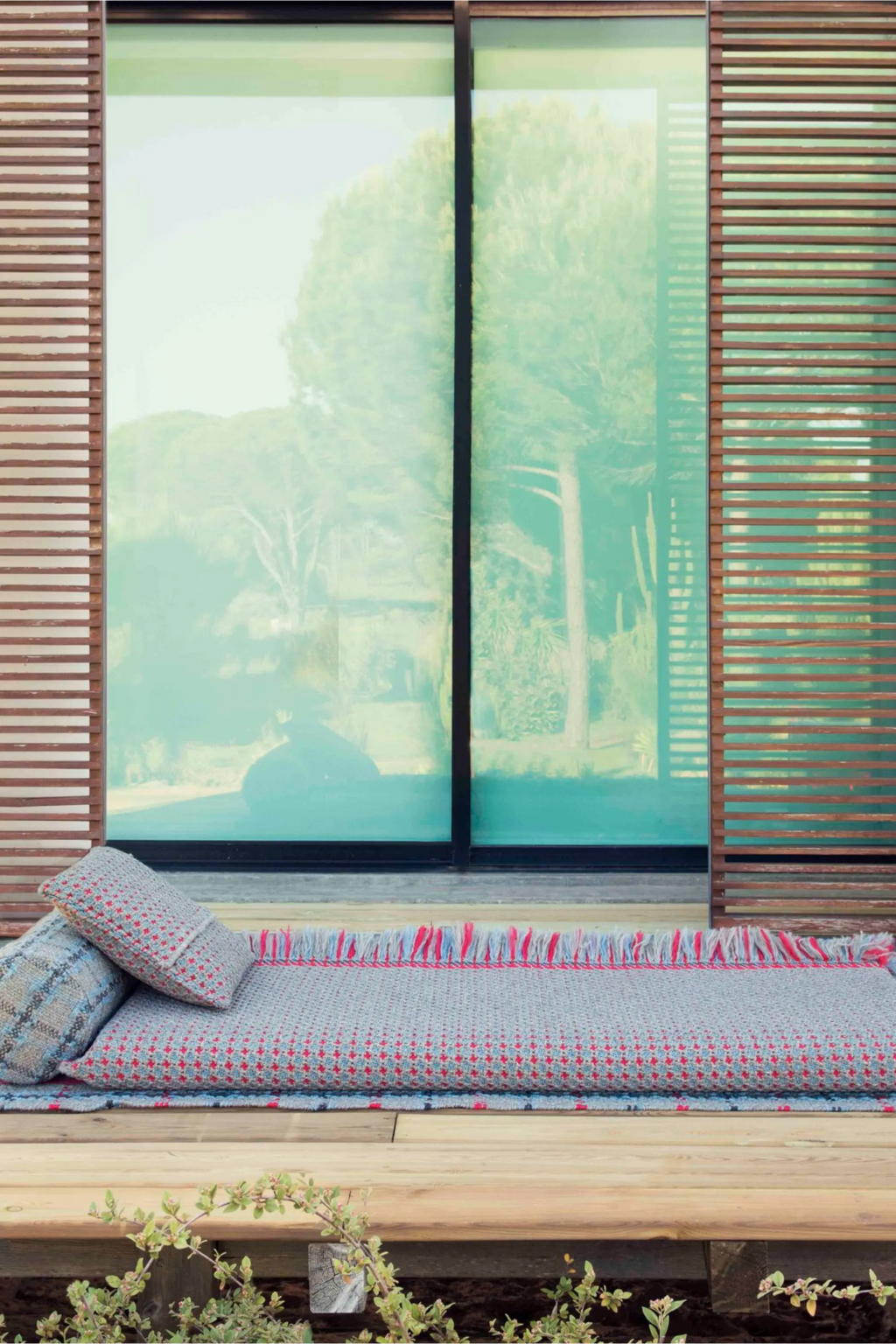 GAN Rugs Garden Layers Composition 06, a modern outdoor sofa set featuring a long floor mattress and accent pillows in a light grey woven fabric with a subtle red and blue geometric pattern. The weather-resistant outdoor cushions are adorned with a playful red and blue fringe along the edge, creating a relaxed, boho-chic aesthetic.