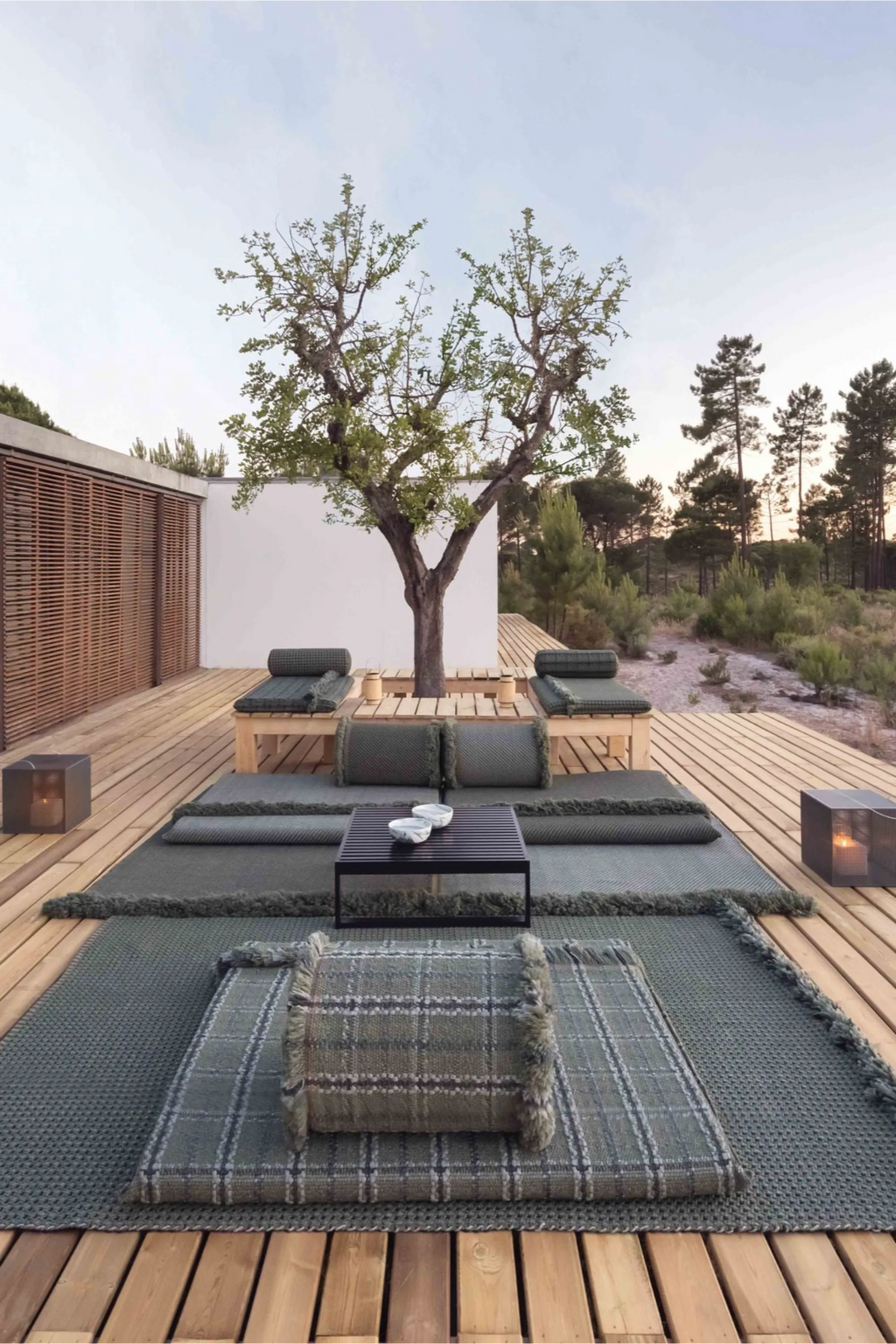 GAN Rugs Garden Layers Composition 04, a luxurious organic modern outdoor lounge set featuring deep olive green and grey-green textured outdoor rugs with fringed edges, layered to create a plush, inviting foundation. This versatile outdoor seating arrangement includes matching woven outdoor cushions, cylindrical bolsters, natural wood daybeds, and a sleek matte black slatted metal coffee table. Ideal for transforming a patio, deck, or garden into a cozy, rustic Mediterranean outdoor living space.