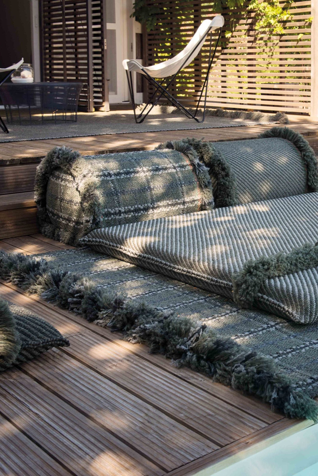 GAN Rugs Garden Layers Composition 03, a modular outdoor sofa featuring woven sage green and grey fabric cushions with distinct striped and plaid patterns, complemented by plush bolster pillows.