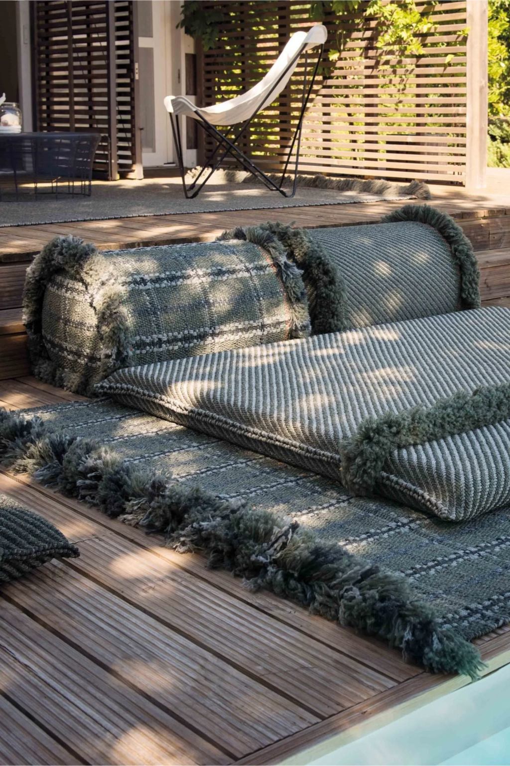 GAN Rugs Garden Layers Composition 04, a modular outdoor sofa featuring hand-woven cushions and bolsters in a natural sage green and grey color palette. This contemporary outdoor lounge set combines striped and plaid patterns with plush, shaggy fringed edges, designed for comfortable relaxation. Ideal for creating a rustic outdoor living space on a patio, deck, or by the pool.