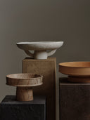 The Dune Raised Bowl made of light brown, veined marble is shown on a dark gray textured plinth on the left. To the right, a smooth orange/terracotta ceramic raised bowl holds a small bunch of red grapes. The background is a solid dark gray.
