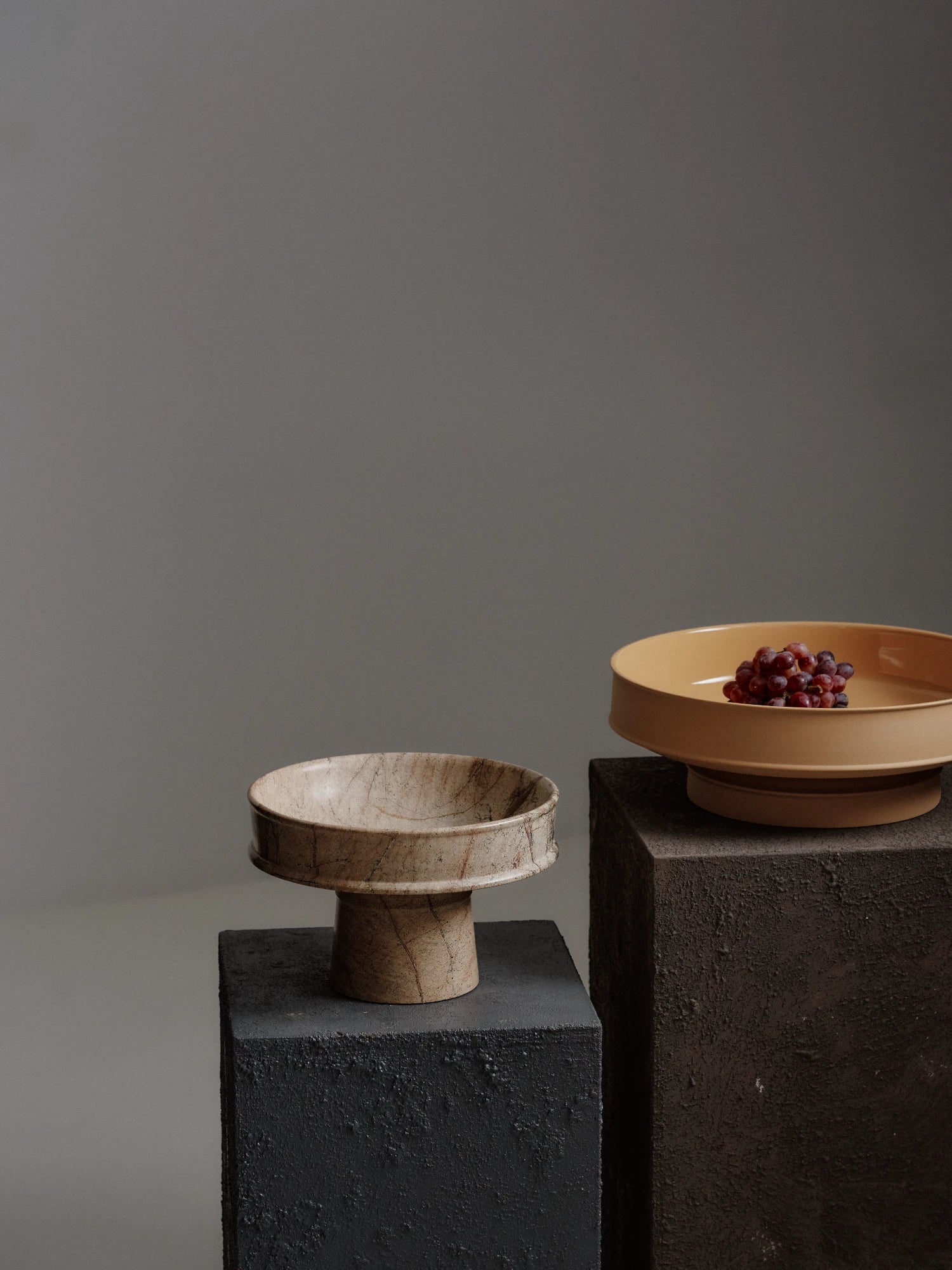 A close-up studio shot of the Dune Raised Bowl made of light brown, veined marble. The image focuses on the bowl's flat, cylindrical pedestal base and the broad, stepped rim of the bowl, which sits on a rough, dark gray plinth.