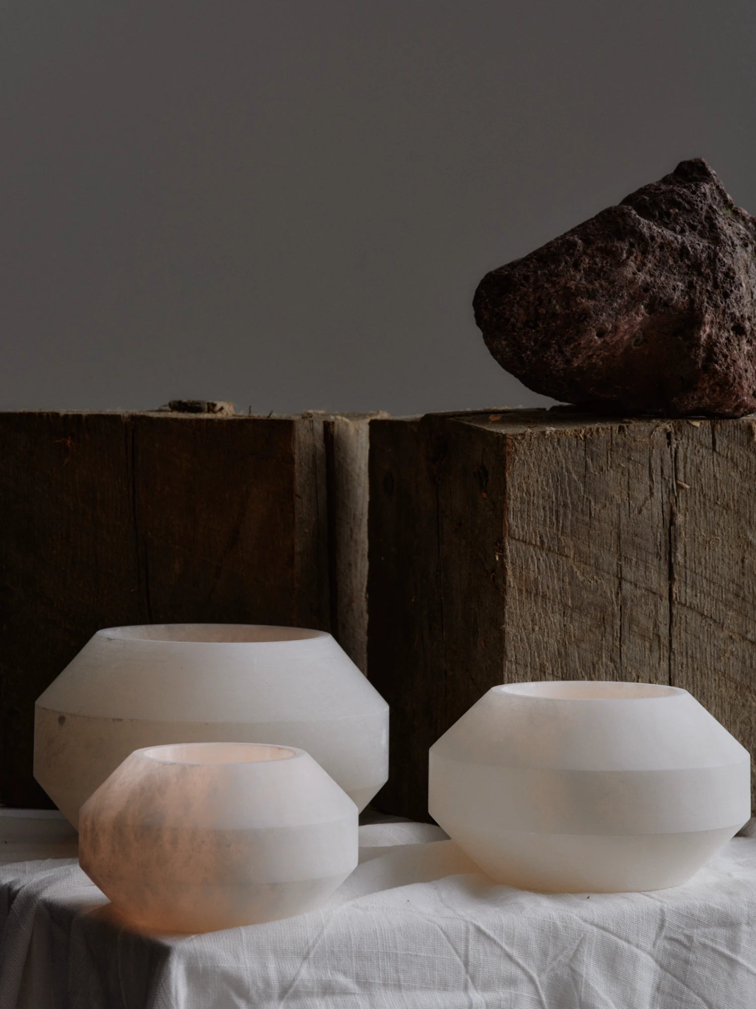 A low-light still-life arrangement featuring three white, translucent alabaster tealight holders on a linen-draped table. The holders are rounded but have distinct horizontal facets. One small holder on the left is lit, showing a warm inner glow, while the two larger holders are unlit. Behind them are two stacked blocks of dark, weathered wood, with a large, reddish-brown piece of volcanic rock resting on top. The background is a solid, dark gray wall.