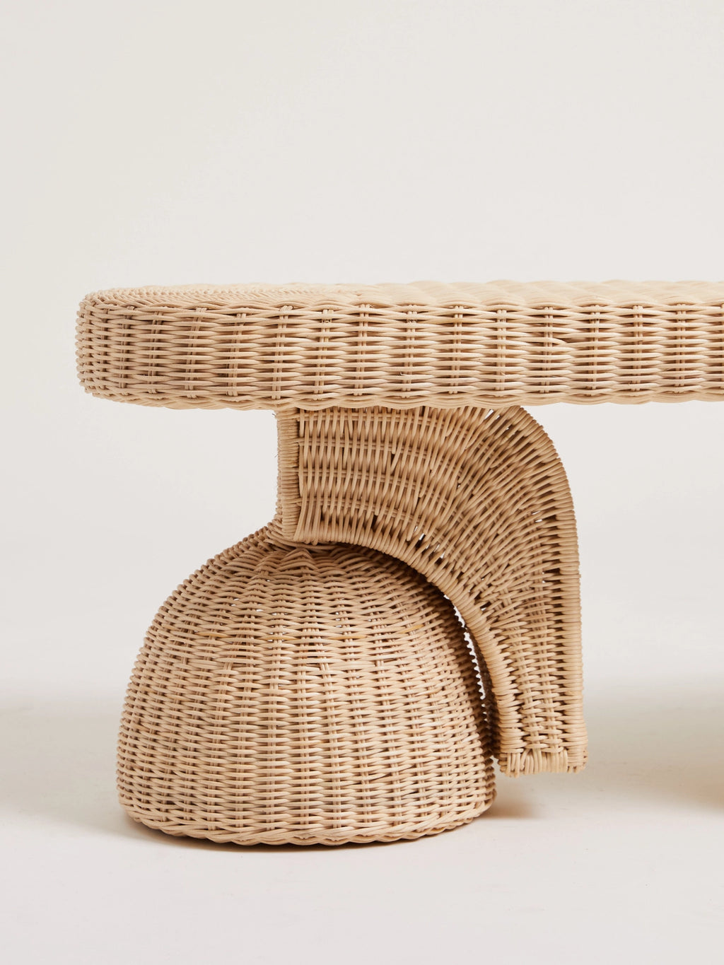 Heaps & Woods Alfred Bench, a natural woven rattan bench featuring a distinctive sculptural and organic design in a light beige finish. This contemporary seating boasts a rounded base and a gracefully curved, C-shaped support leading to an oval top, all crafted from finely woven rattan. Ideal as an entryway bench, bedroom accent, or for organic modern living room decor.