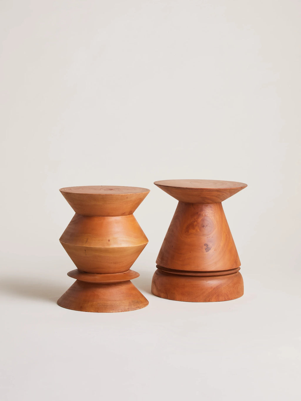 Heaps & Woods Thierry Stool, a pair of sculptural solid wood accent stools in warm natural wood tones with smooth, polished finishes. One stool features a distinctive hourglass-like shape with stacked geometric segments, while the other presents a conical form with a flared top, both embodying mid-century modern and organic design aesthetics. These versatile natural wood stools are perfect as side tables, plant stands, or decorative accents for living rooms, bedrooms, or entryways.