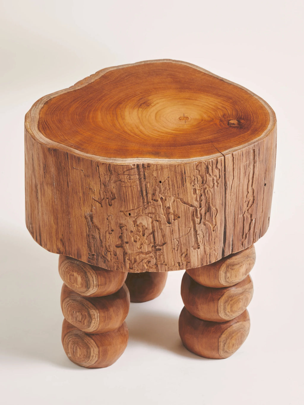 Heaps & Woods Vic Stool, an organic modern solid wood stool crafted from a natural tree stump slice, featuring a warm medium brown live edge top with visible growth rings and an irregular, organic shape. The rustic cylindrical body showcases natural bark texture and intricate wormhole patterns, supported by three chunky, stacked rounded wood legs. This handcrafted wooden stool is perfect as a unique side table or accent piece for wabi-sabi, organic modern, or rustic living rooms and bedrooms.
