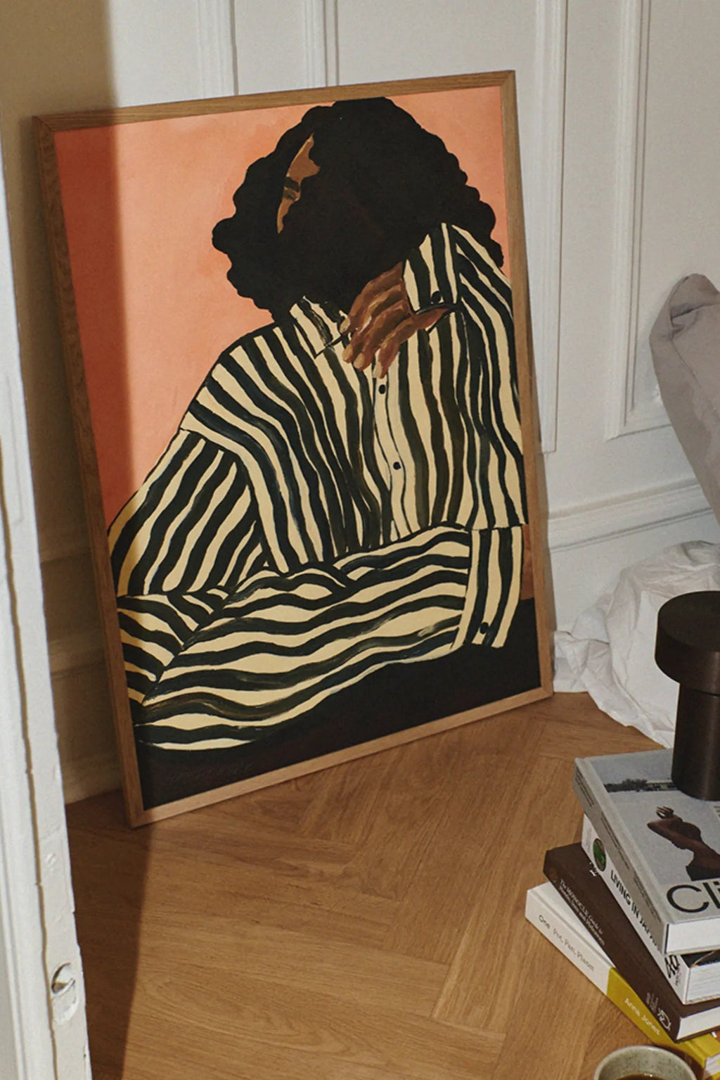 The Poster Club Serene Stripes, a contemporary abstract portrait art print featuring a figure in a bold black and cream striped shirt, with flowing black hair and warm brown skin tones, set against a soft peachy pink background. This minimalist and graphic wall art, presented in a natural oak frame, is ideal for modern living room decor, a stylish bedroom accent, or as a striking addition to a gallery wall.