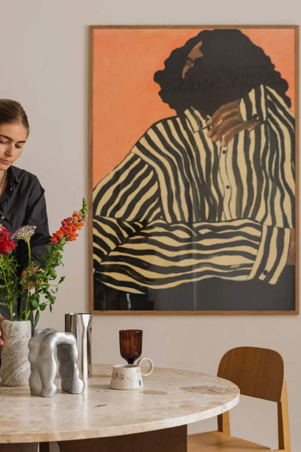 The Poster Club Serene Stripes, a contemporary abstract figurative art print featuring a bold graphic portrait of a person in a black and beige striped shirt against a vibrant peachy-orange background, framed in a light natural oak frame. This unique framed poster is perfect as modern wall art for a statement gallery wall, living room, or bedroom decor.