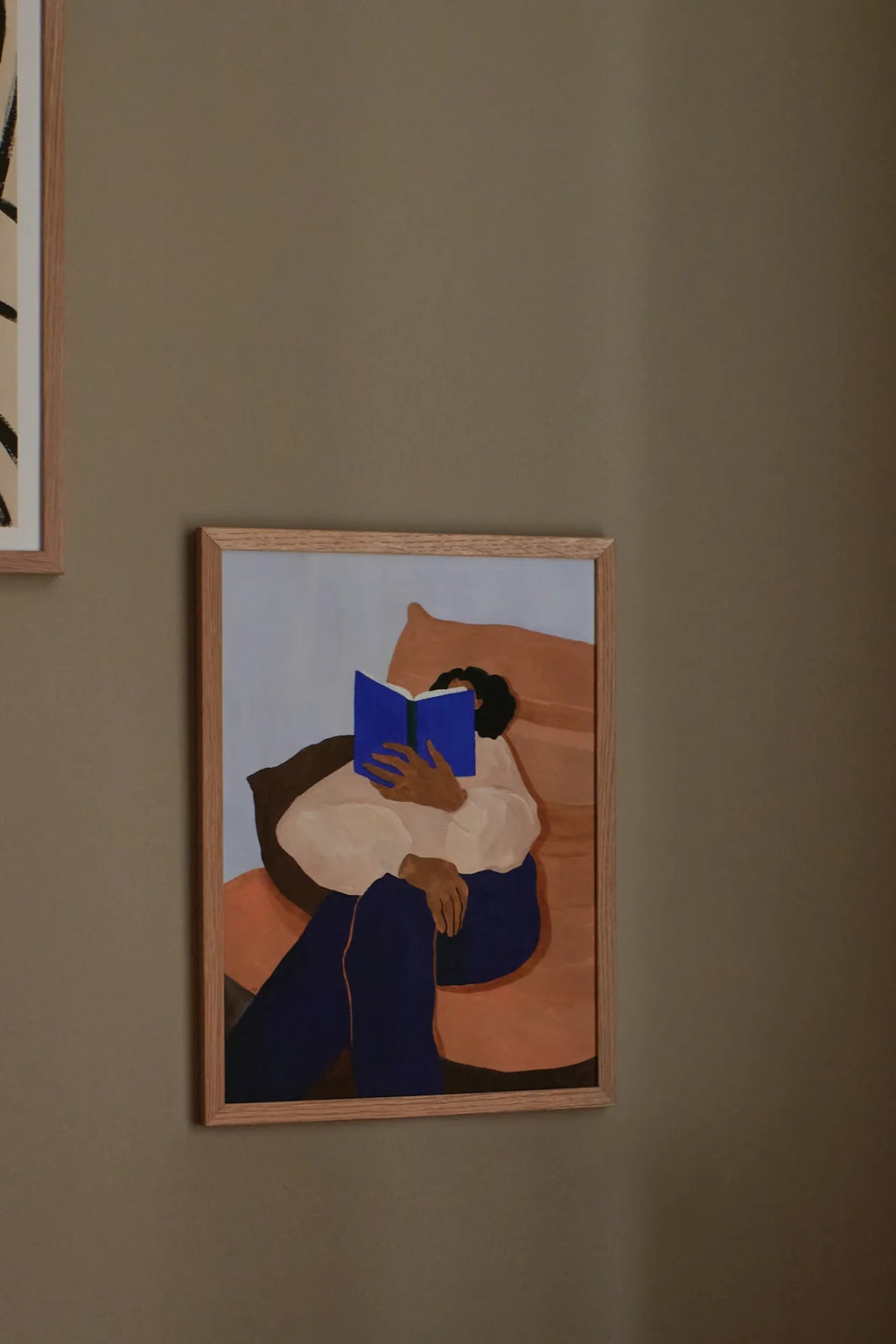 The Poster Club The Togo, an abstract minimalist art print depicting a person reading a bright blue book, dressed in a cream top and dark blue pants, seated on a terracotta cushion against a muted blue background. This contemporary wall art is framed in a light natural oak wood frame, ideal for modern living rooms, minimalist bedrooms, or an organic modern gallery wall.