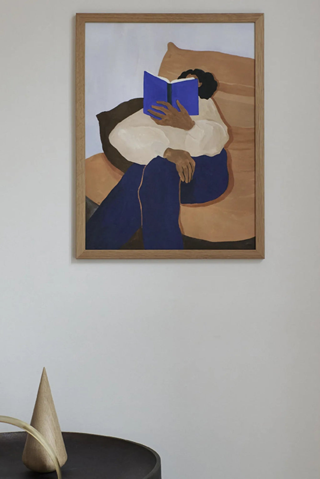 The Poster Club The Togo, an abstract figurative art print depicting a person seated and engrossed in a vibrant blue book, rendered in rich navy blue, off-white, and warm brown tones against a soft light blue background. This contemporary wall art is presented in a natural oak frame with a clean white mat. Ideal for minimalist living rooms, cozy reading nooks, or modern bedroom decor.