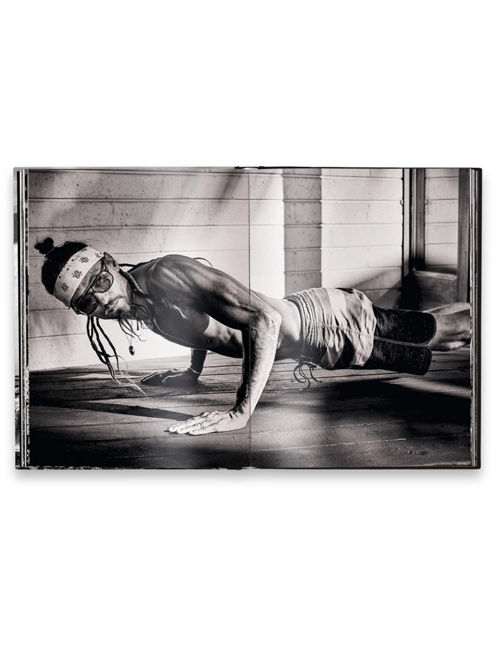 Book Hawaii, an open photography book displaying a black and white image of a man with dreadlocks and sunglasses doing a push-up on a wooden floor, wearing a bandana and striped shorts. This captivating art book is perfect for photography enthusiasts and those interested in Hawaiian culture and lifestyle.