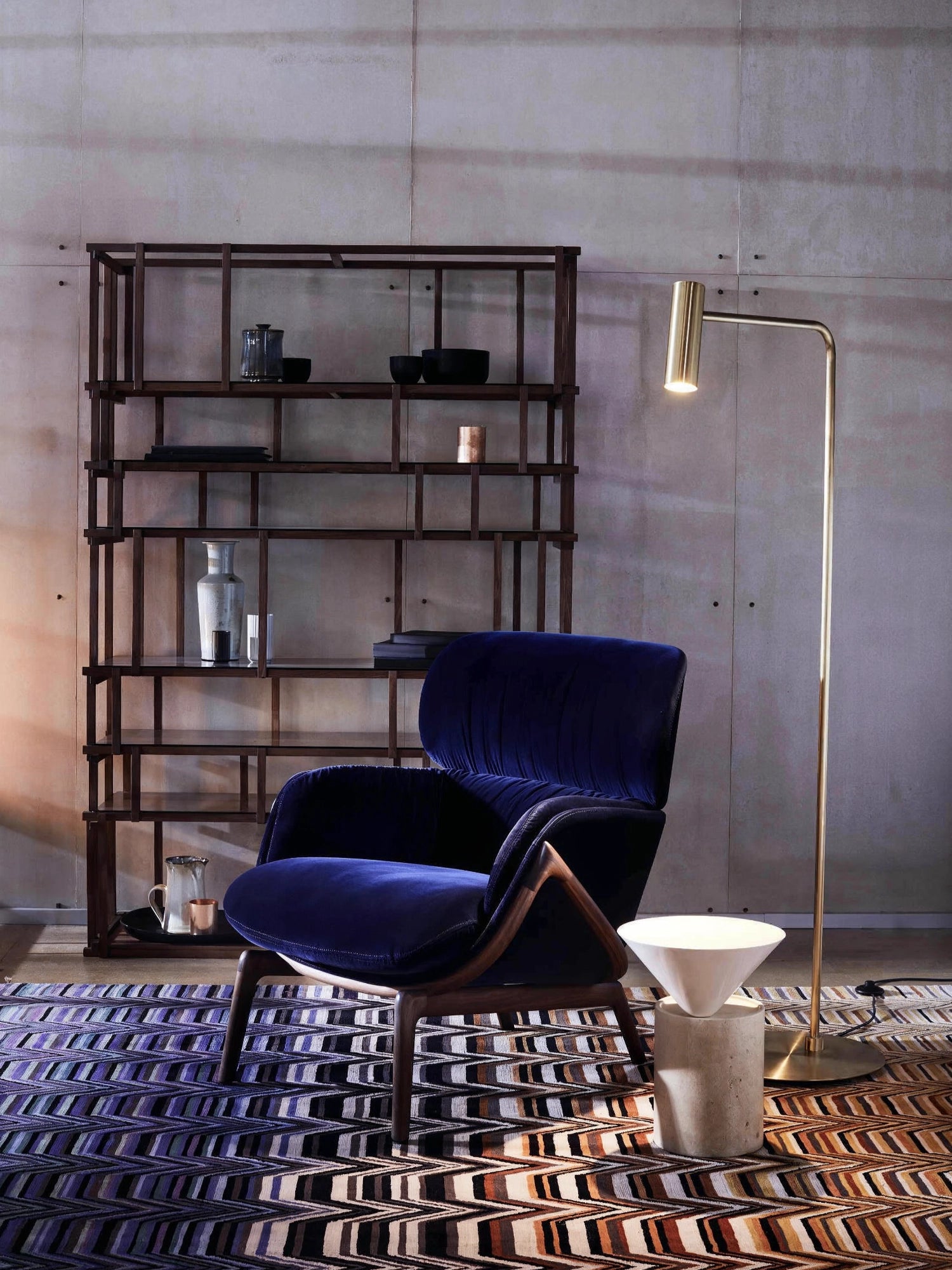 CTO Lighting Heron Floor Lamp, a luxurious brass floor lamp with a cylindrical head, providing focused illumination in a stylish room with a blue velvet armchair and wooden shelving.