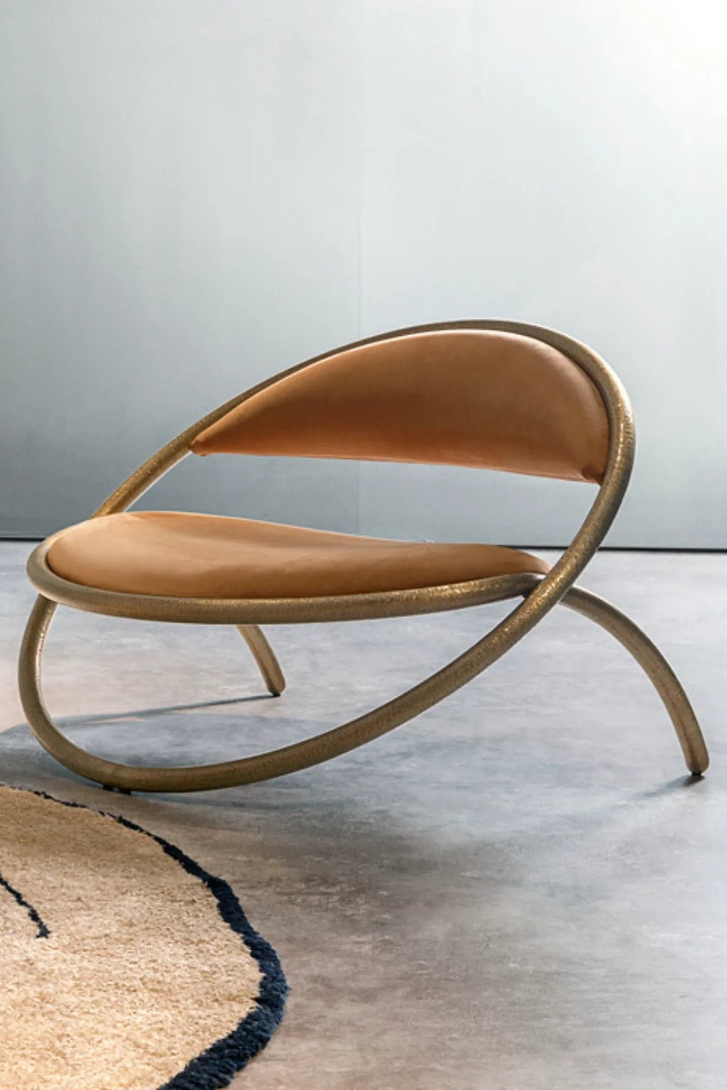 Hessentia Ryo Armchair, a sculptural organic modern lounge chair featuring a continuous, curved frame in textured, warm bronze-toned metal, cradling a crescent-shaped seat and backrest upholstered in smooth camel-toned leather. This luxurious contemporary accent chair is ideal for sophisticated living rooms, hotel lobbies, or modern bedroom spaces.