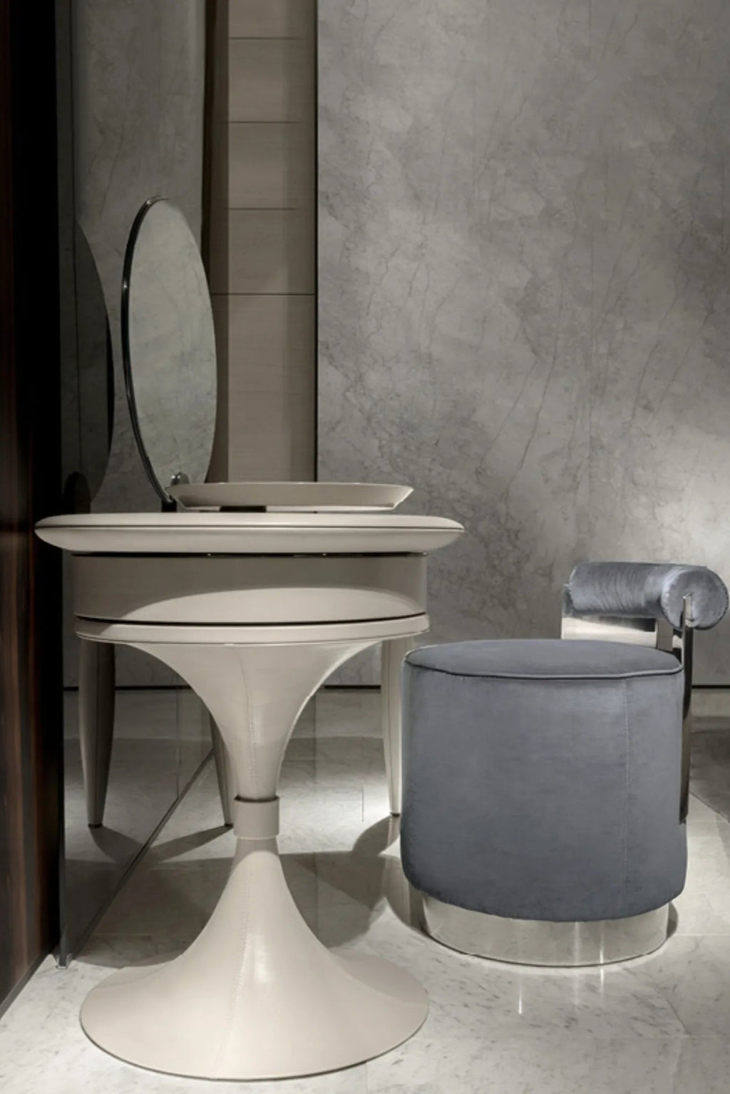 Hessentia Shelly Pouf, a sophisticated grey velvet cylinder pouf featuring plush velvet upholstery with subtle piping detail and a sleek polished chrome base. Its clean, minimalist design and elegant contemporary aesthetic make it a versatile luxury accent piece. Perfect as a modern vanity stool for a chic dressing table or as stylish accent seating in a living room or bedroom.