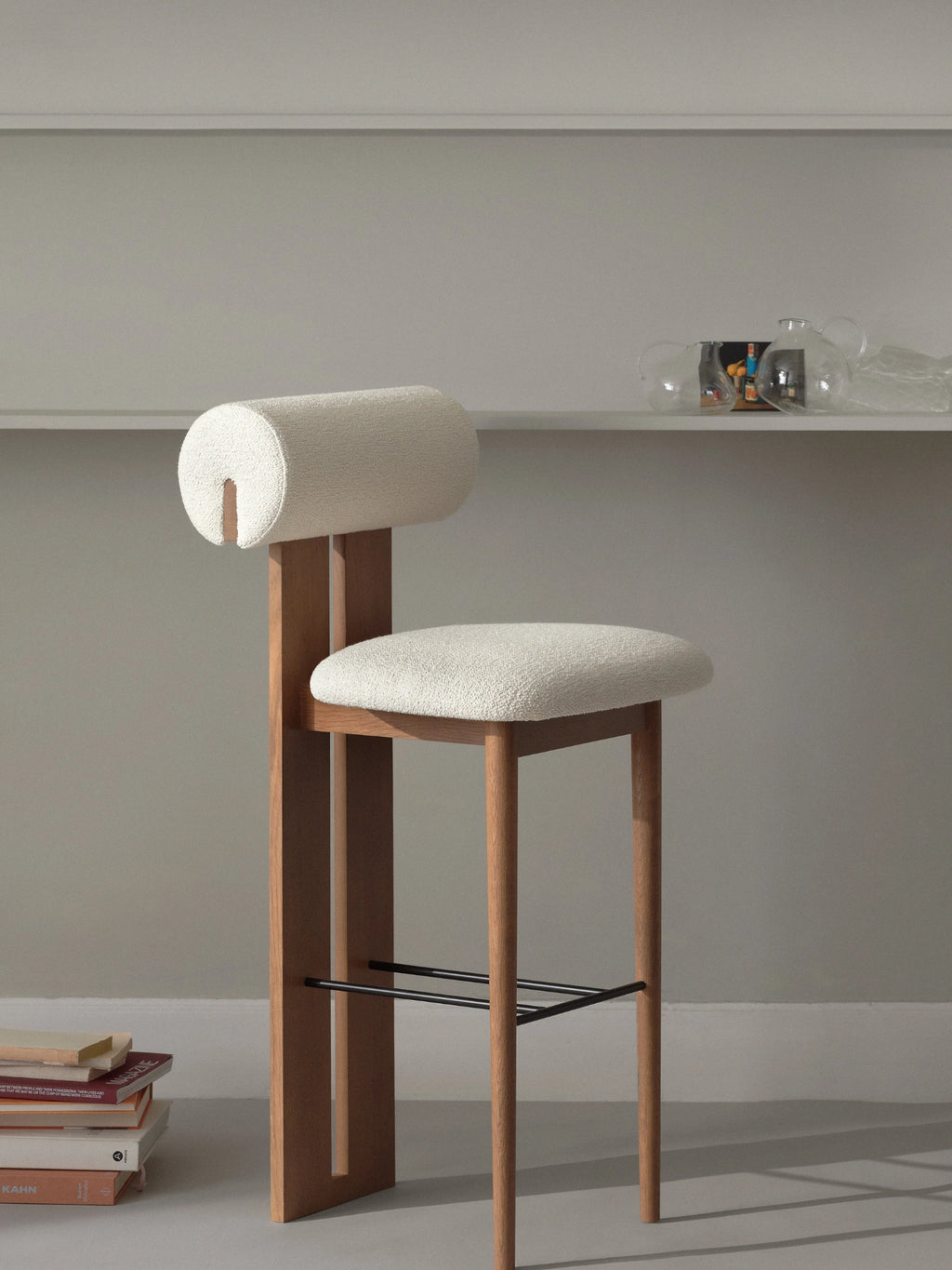NORR11 Hippo Bar Chair, a contemporary bar stool featuring a sturdy solid oak wood frame with slender tapered legs and a distinctive rectangular back support. It boasts a plush rounded seat and a cylindrical backrest, both upholstered in a textured cream bouclé fabric, complemented by a matte black metal footrest. This organic modern bar chair is perfect for Scandinavian kitchen islands or minimalist home bar areas.