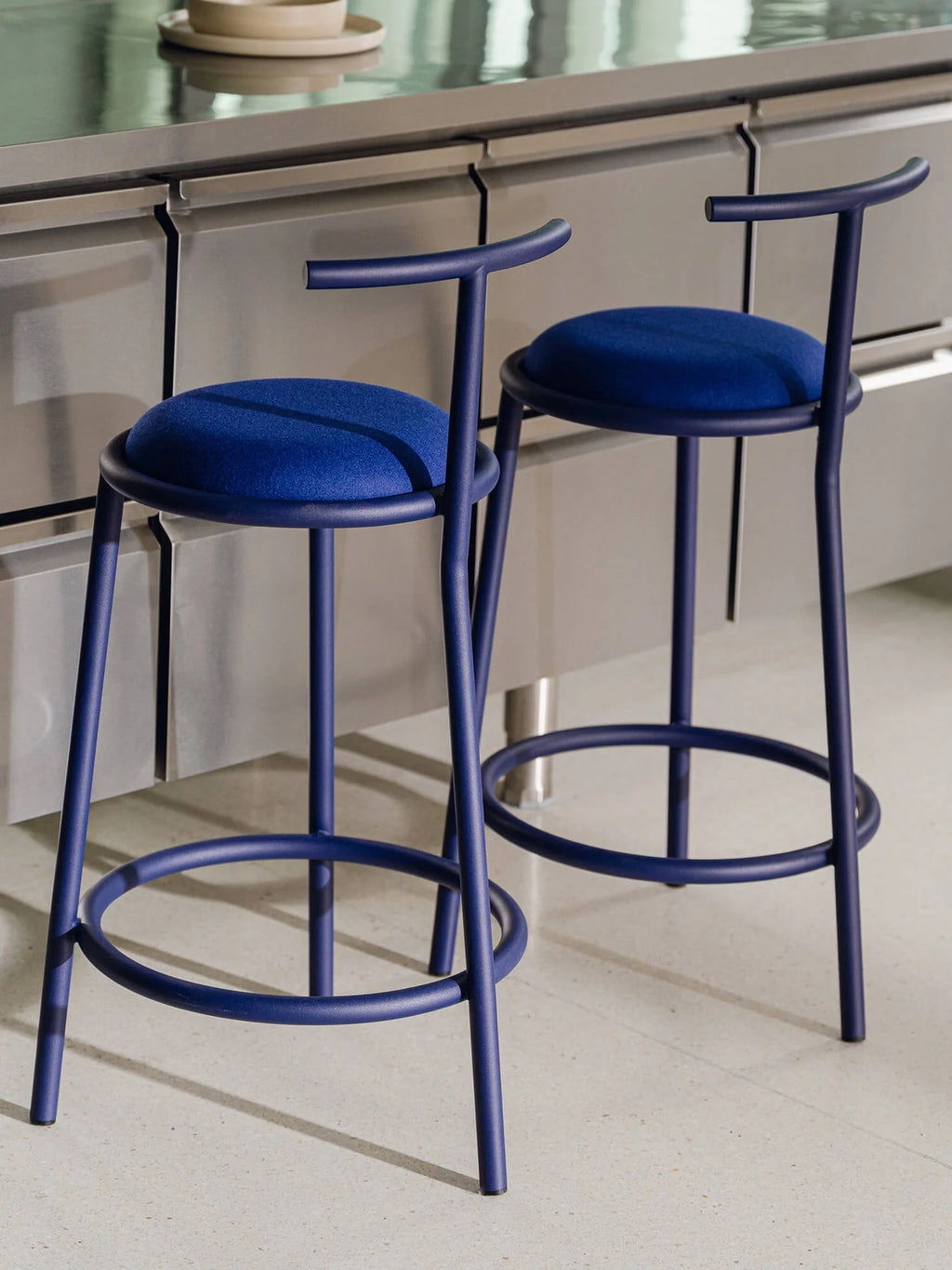 noo.ma Hogo Bar Stool w Backrest, a modern minimalist bar stool featuring a sleek matte blue powder-coated metal frame with a circular footrest and a gently curved back.