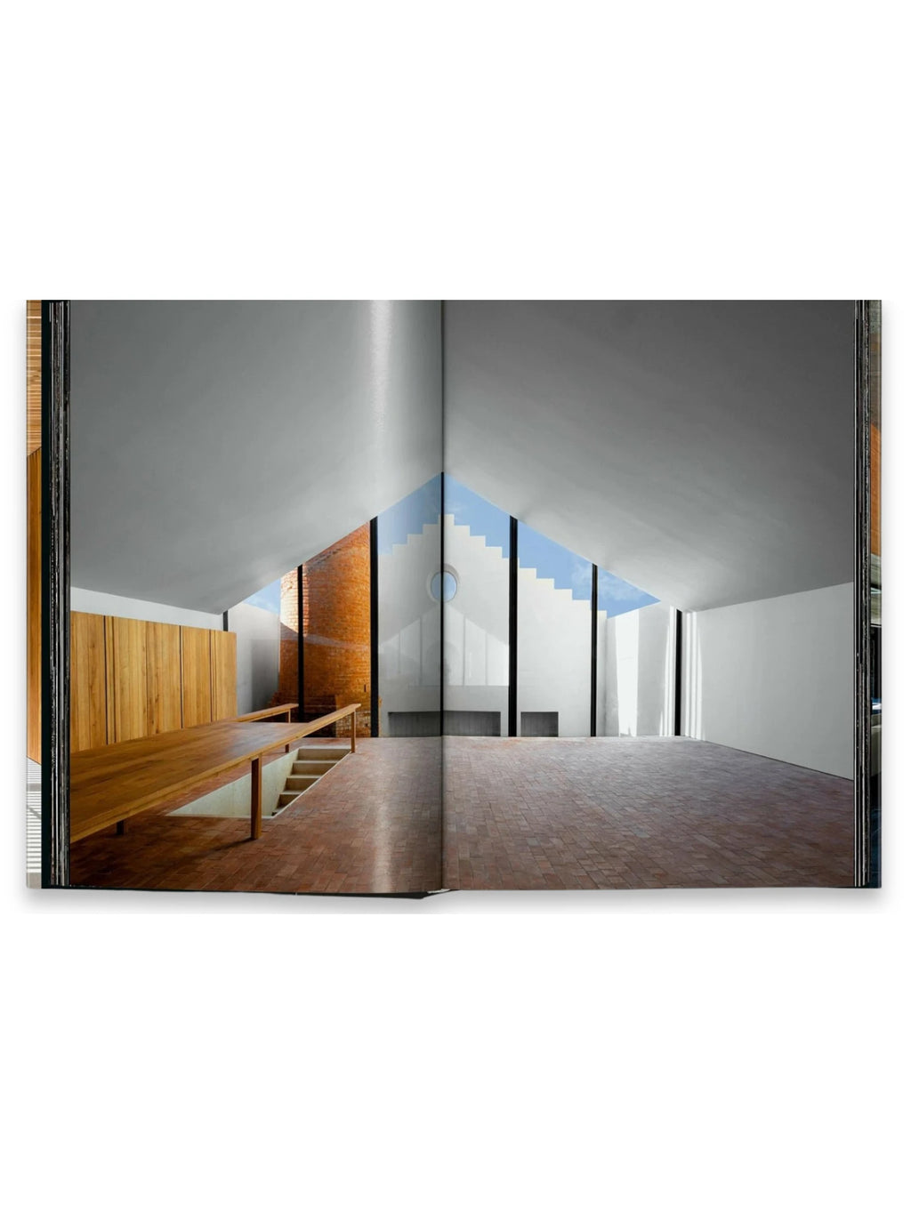 Book Homes for Our Time. Vol. 2, an open architectural book displaying a minimalist, modern interior with vaulted white ceilings, a long natural wood built-in desk, exposed brick wall accents, and an expansive window revealing a clear blue sky. This captivating architecture book is ideal for design professionals, students, and enthusiasts seeking inspiration for contemporary living spaces.
