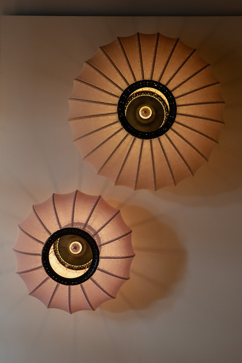Made by Hand Knit-Wit Ø76 Oval Wall Light, a contemporary textile wall light featuring a soft blush pink knitted yarn shade with a distinctive pleated texture and a dark central ring, emitting a warm ambient glow. Perfect for Scandinavian living rooms, as a decorative bedroom accent light, or for creating soft lighting in modern interiors.