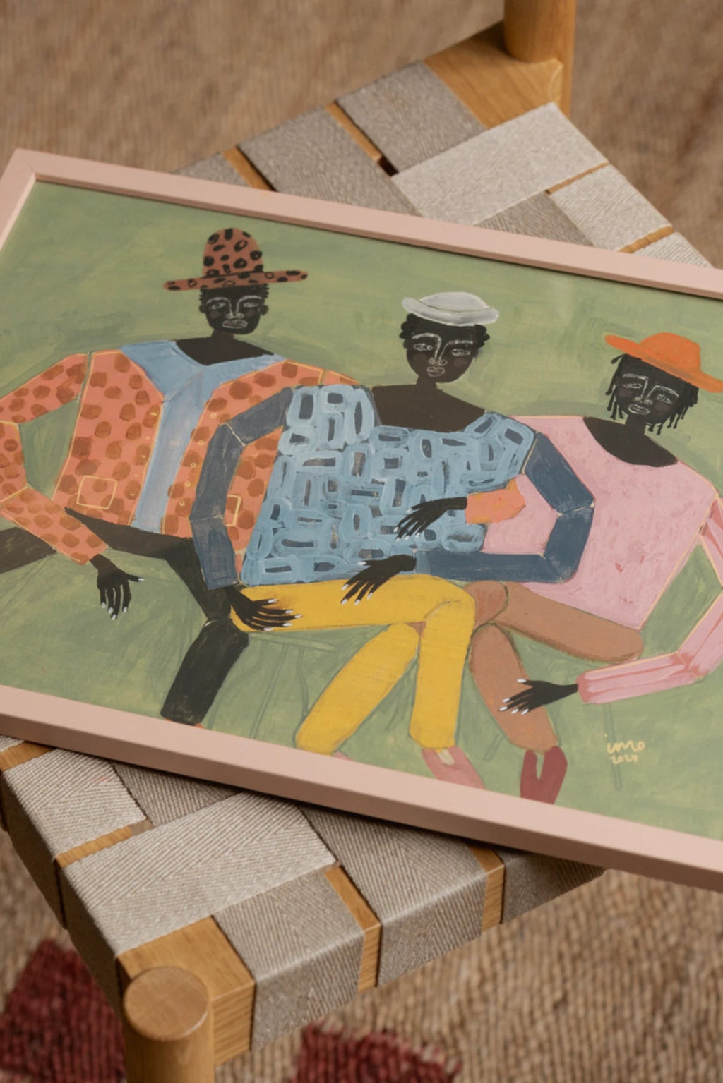 The Poster Club Hanging Out, a colorful contemporary folk art print depicting three abstract figures in vibrant clothing and distinct hats against a pastel green background, showcasing expressive brushstrokes and a painterly texture. This framed art print is presented in a light pink wood frame, enhancing its playful and modern aesthetic. It is ideal as a statement piece of wall art for a living room, bedroom, or a creative gallery wall.