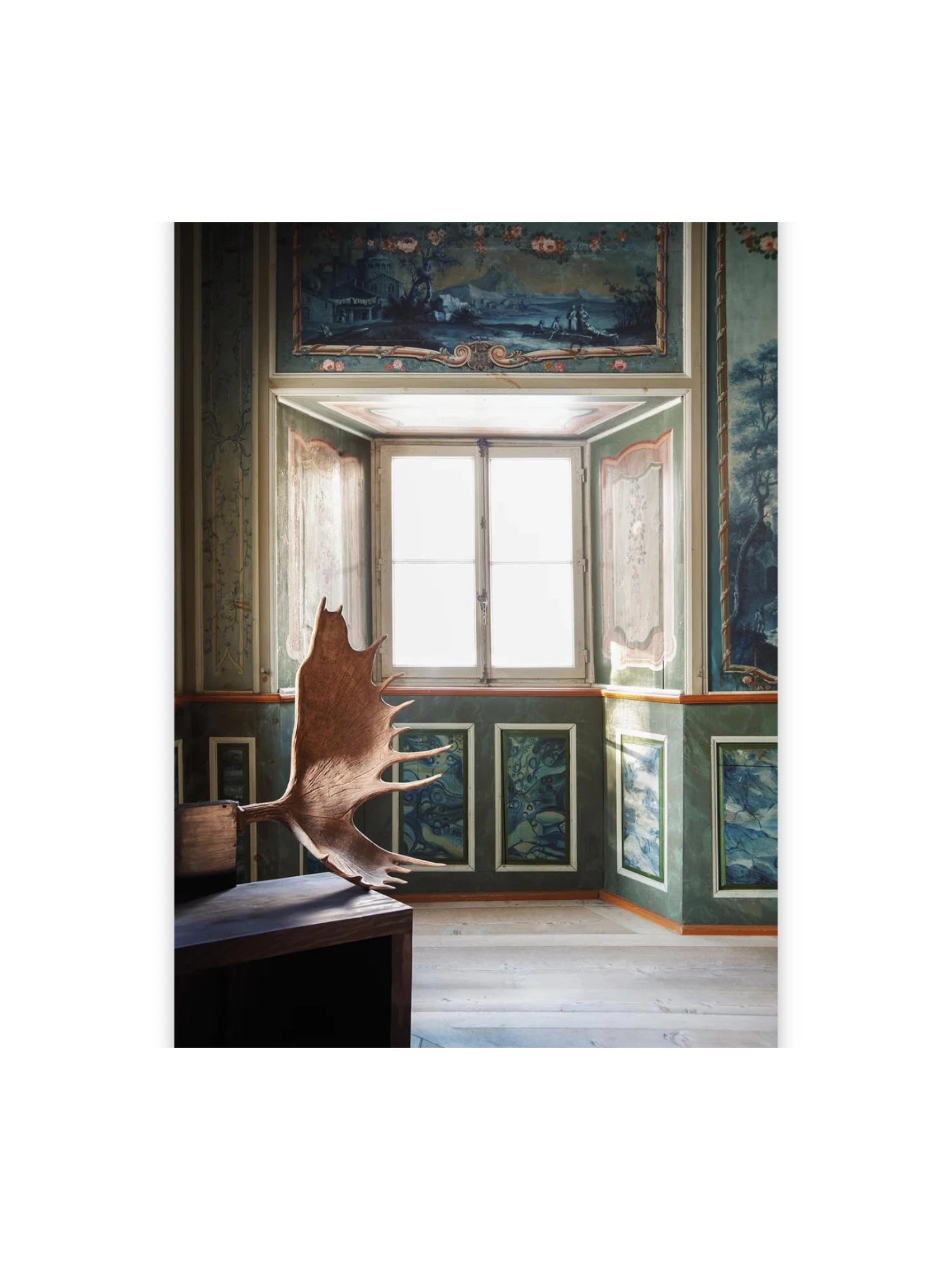 Book In Focus, a captivating photography book showcasing a large, rustic moose antler resting on a dark wooden table in a lavish room with highly detailed, ornate green and blue painted murals. This art book is perfect for collectors of fine art photography and those interested in historical interiors.