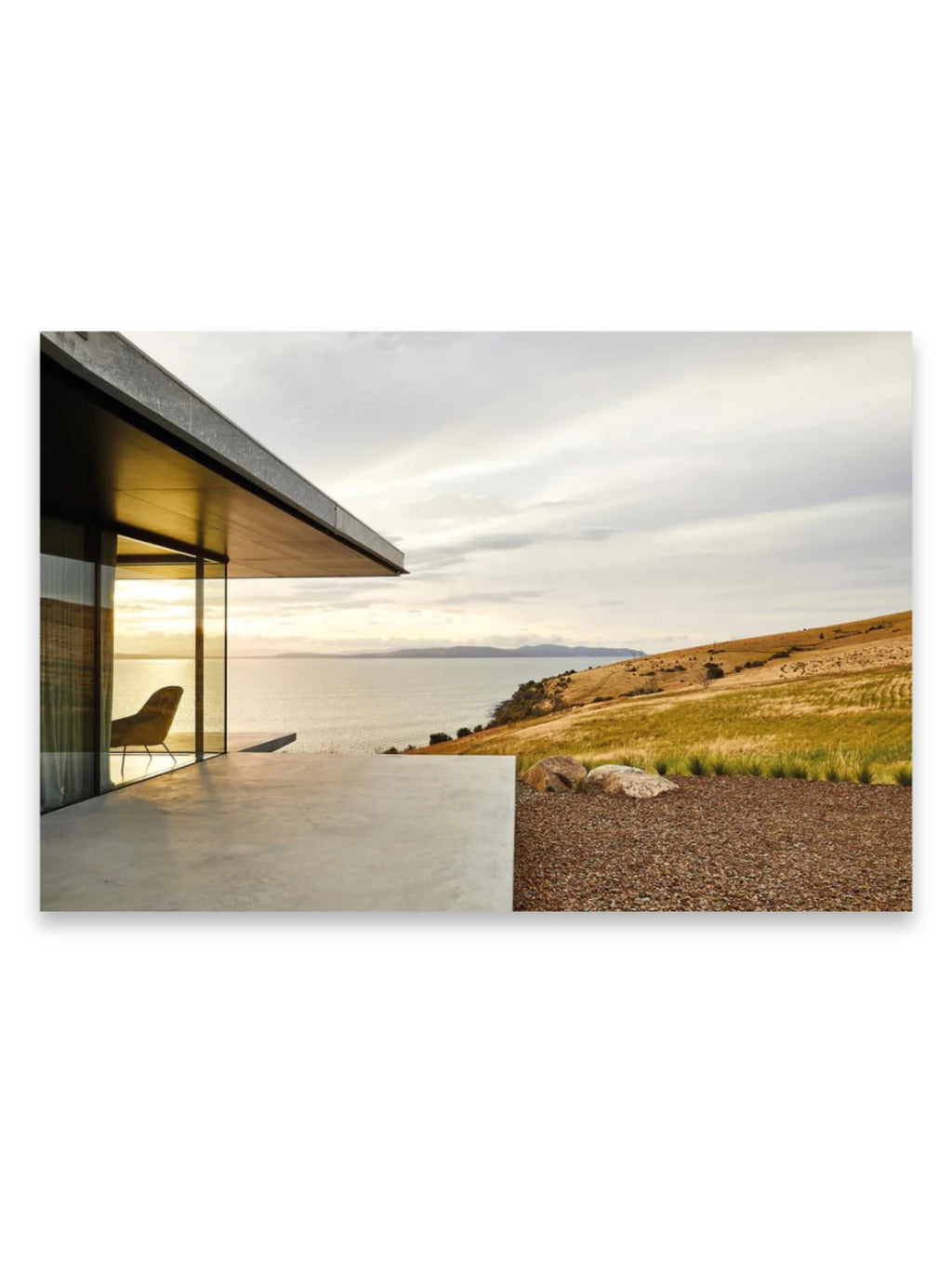 Book In Focus, a modern luxury architectural design book showcasing a sleek home with expansive glass walls, a concrete patio, and a minimalist interior chair overlooking a scenic ocean and grassy hillside at sunset. This premium book is ideal for architects, interior designers, and luxury home enthusiasts seeking inspiration.
