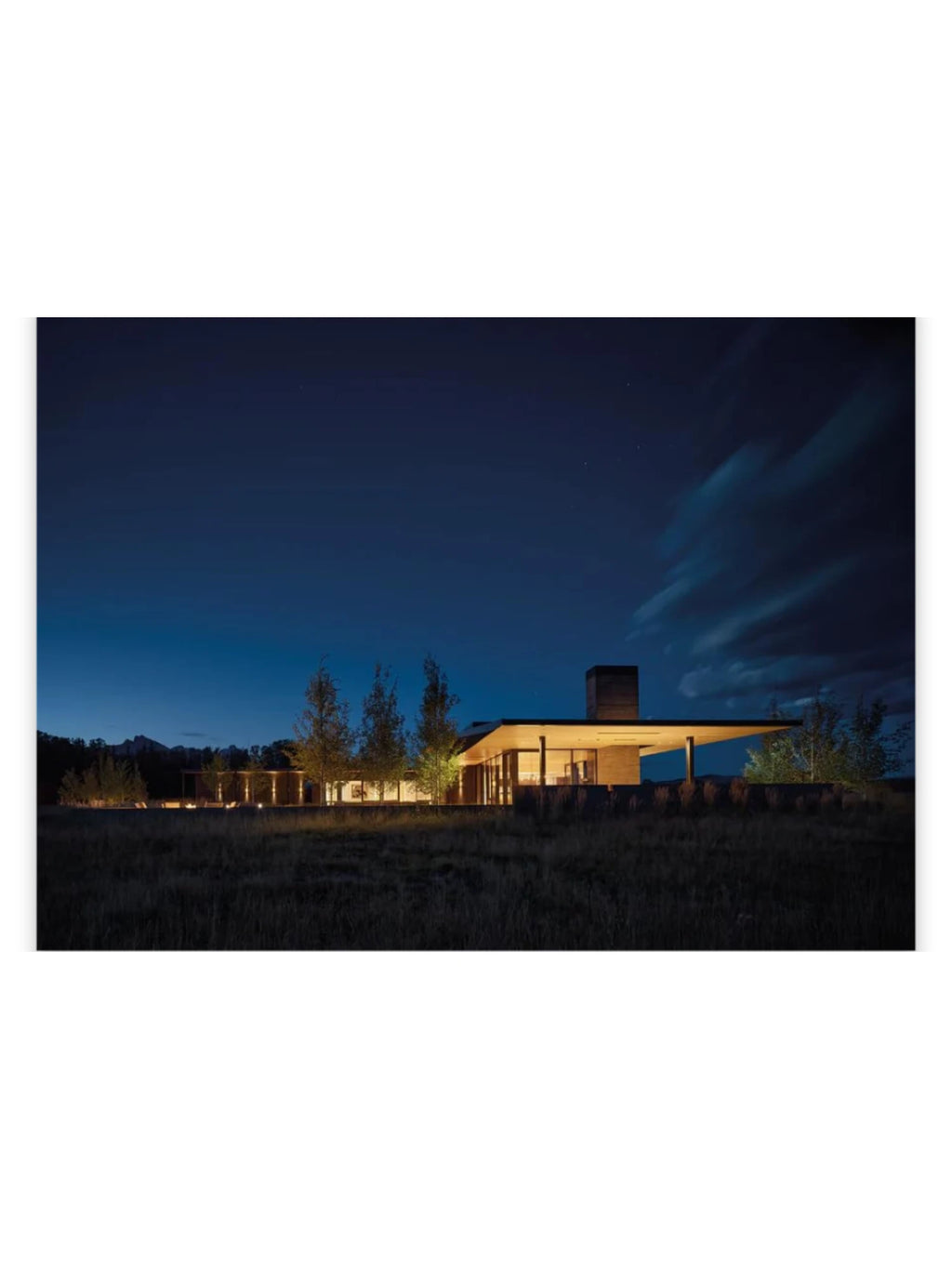 Book In Focus, a modern luxury home with warm interior lighting against a dark blue twilight sky and dry grass landscape. This architecture book offers inspiration for contemporary home design enthusiasts.