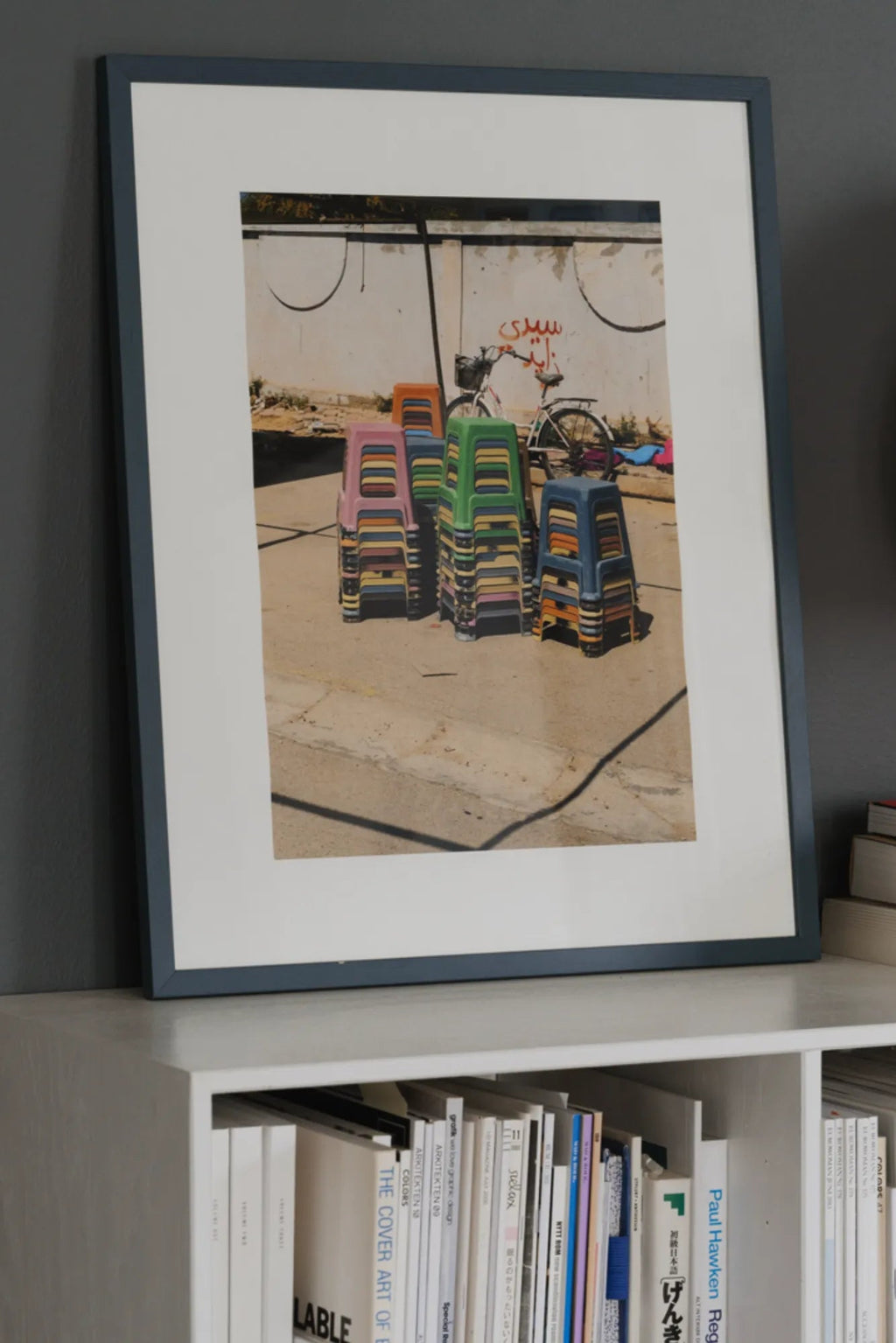 The Poster Club The Souk, a vibrant street photography art print showcasing stacks of colorful plastic stools in muted pink, green, orange, and blue hues, set against an urban wall with a bicycle. This contemporary poster is elegantly framed with a crisp white mat and a sleek dark grey-blue frame. Ideal as eye-catching wall art for a modern living room, bedroom, or a dynamic gallery wall in urban-inspired interiors.