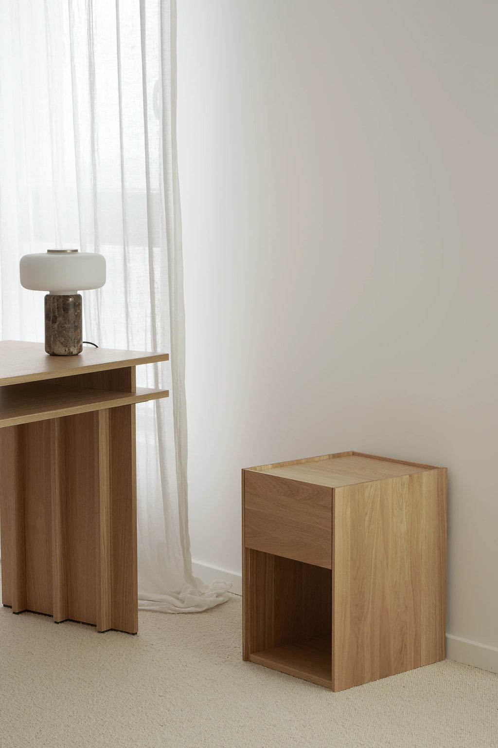 TAMO Kari Nightstand, an elegant light brown oak wooden nightstand with one drawer, an open shelf, and raised lip tabletop, displayed in a minimalist room. Ideal for modern bedroom storage.