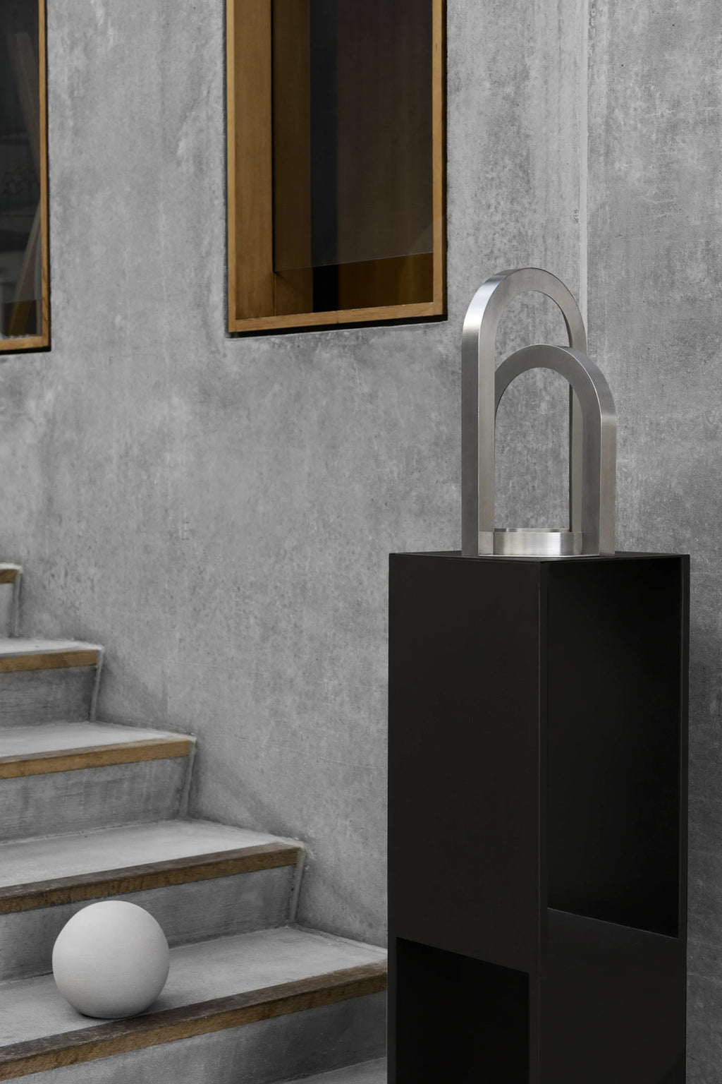 Kristina Dam Arch Sculpture, a striking modern minimalist sculpture crafted from brushed stainless steel, featuring two nested, rounded geometric arch shapes rising from a cylindrical base. This contemporary metal art piece offers a sleek, architectural design in a silver finish, perfect for adding an industrial chic or brutalist touch to modern shelf styling, a living room console, or home office decor.