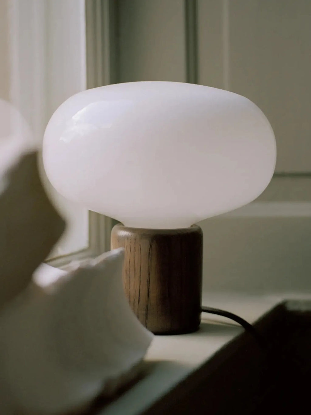 New Works Karl-Johan Table Lamp, showcasing a premium white milky blown glass shade atop a dark stained oak cylindrical base. A minimalist, luxury design for contemporary living spaces.