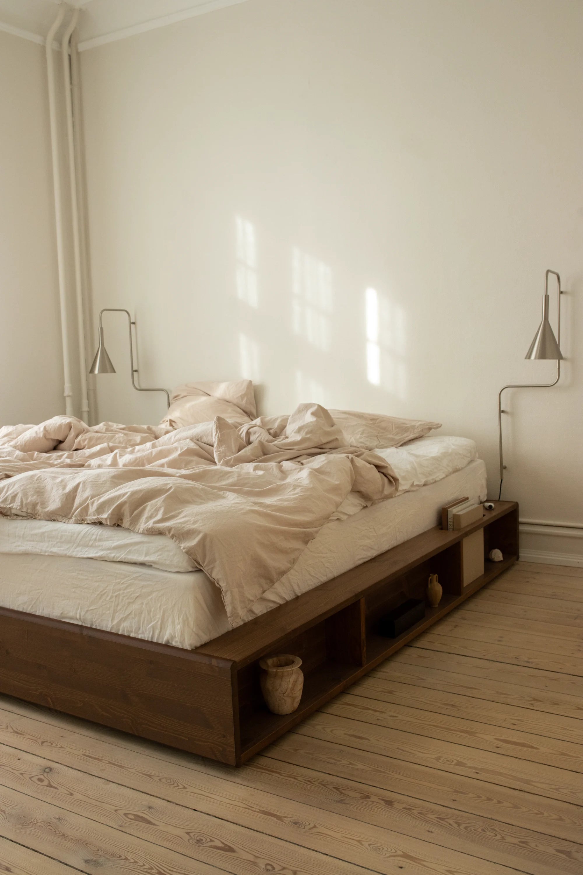 Wooden bed with beige bedding in a minimalistic room with light wood flooring and white walls.