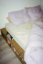 Bed with white bedding and purple pillows against a plain wall.