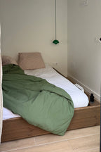 Bed with green blanket and white sheets in a room with beige walls and wooden floor.
