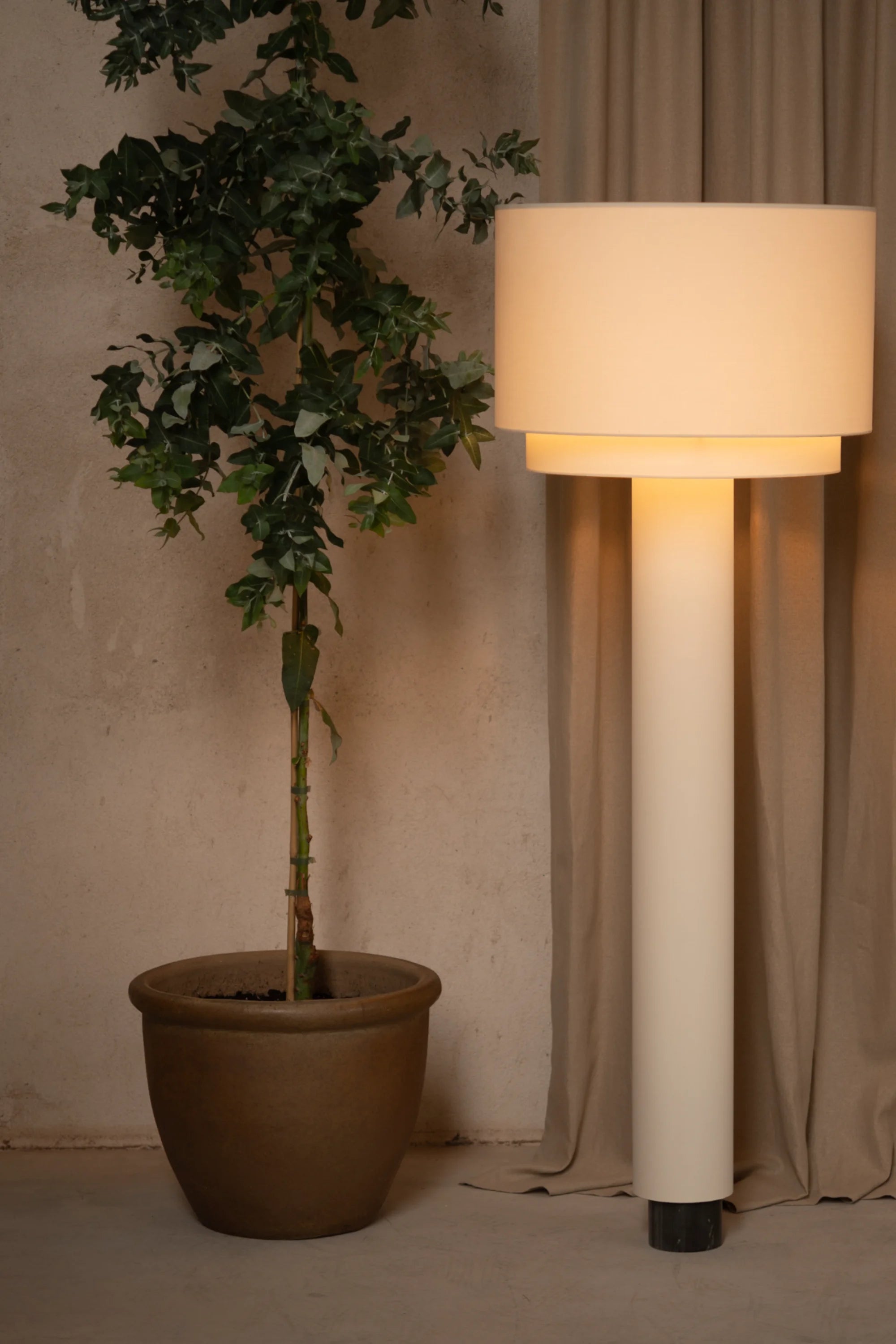 Tall floor lamp with a beige shade next to a potted plant against a beige wall.