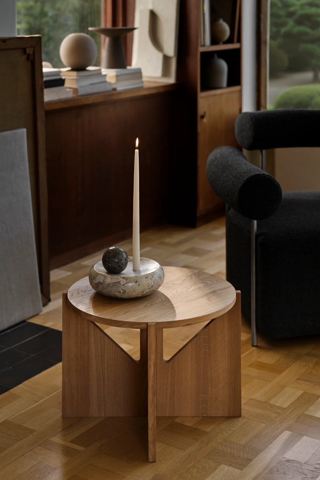 Kristina Dam Coherent Candle Sculpture, an organic modern candle holder featuring a rounded, bowl-shaped base crafted from light, porous travertine stone, topped with a smooth white marble disc. It elegantly holds an off-white tapered candle and a dark, textured stone sphere, creating a balanced sculptural arrangement. This minimalist piece is ideal for sophisticated shelf styling or as a contemporary dining table centerpiece.