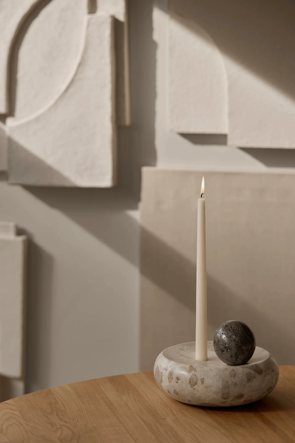 Kristina Dam Coherent Candle Sculpture, a minimalist organic modern candle holder crafted from natural beige travertine stone with a rounded, sculptural base designed for a single cream taper candle. It features a separate dark grey textured stone sphere, creating a contemporary decorative object with brutalist influences. Perfect for sophisticated shelf styling or as a modern dining table centerpiece.