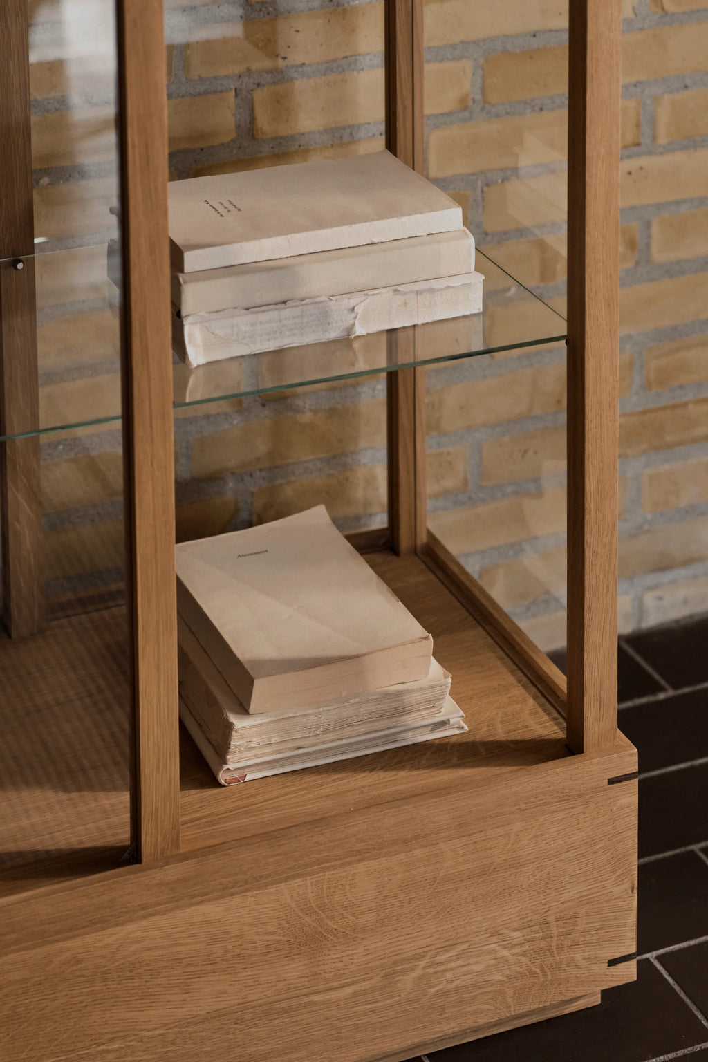 Kristina Dam Japanese Cabinet L, a tall minimalist display cabinet crafted from natural solid oak with a smooth wood grain and clean, architectural lines inspired by Japanese design. This contemporary storage unit features clear glass shelves and a sturdy oak base, showcasing exquisite exposed joinery details. Ideal for elegant shelf styling and organized storage in Scandinavian living rooms, modern dining spaces, or minimalist home offices.