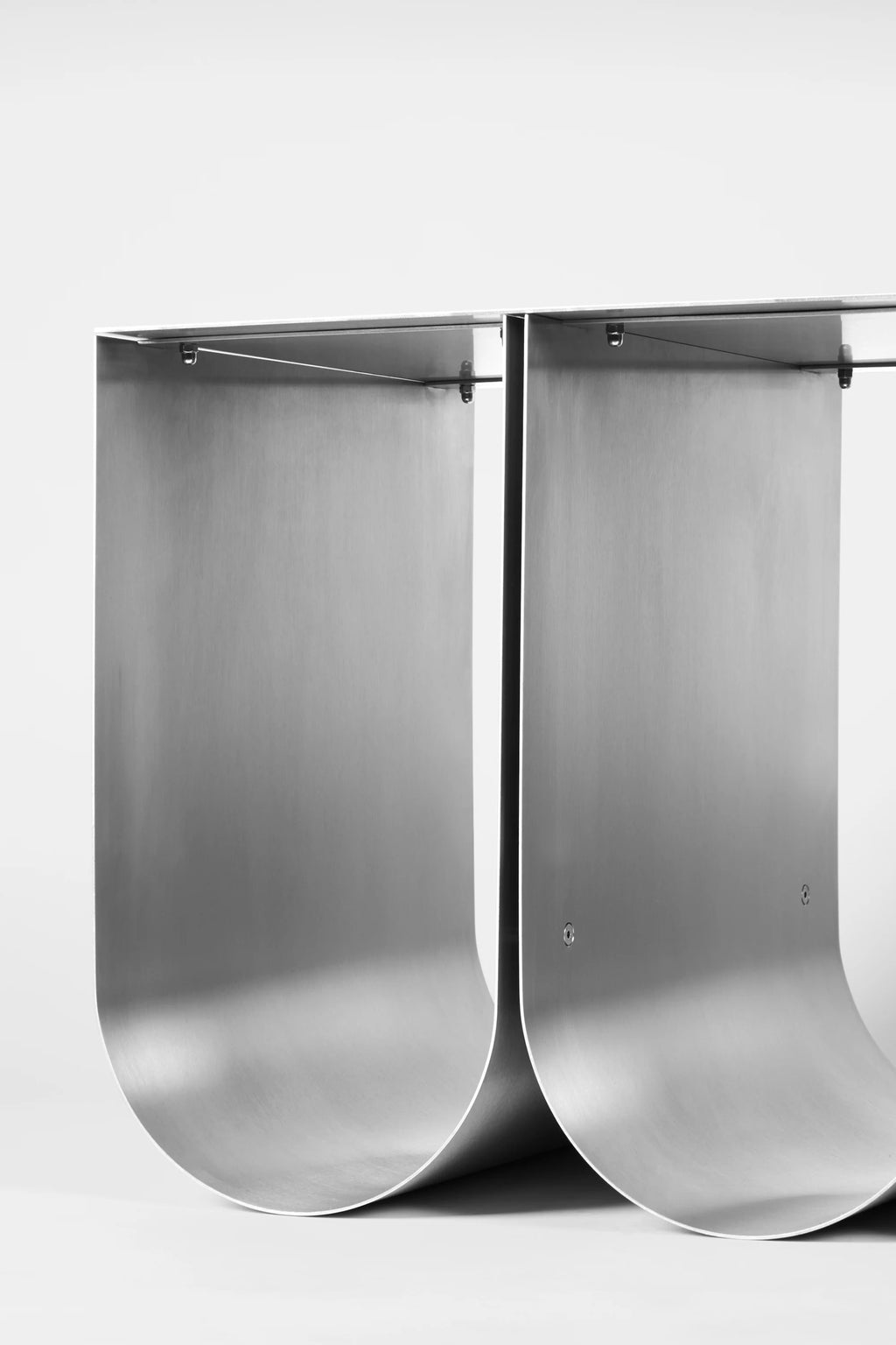 Kristina Dam Curved Bench, a striking modern minimalist bench crafted from brushed stainless steel, featuring two bold, curved silver metal sheets that form a sculptural base supporting a sleek, flat top surface. Its industrial chic aesthetic and clean lines embody contemporary design, making it a perfect entryway bench or living room accent for minimalist and brutalist interiors.
