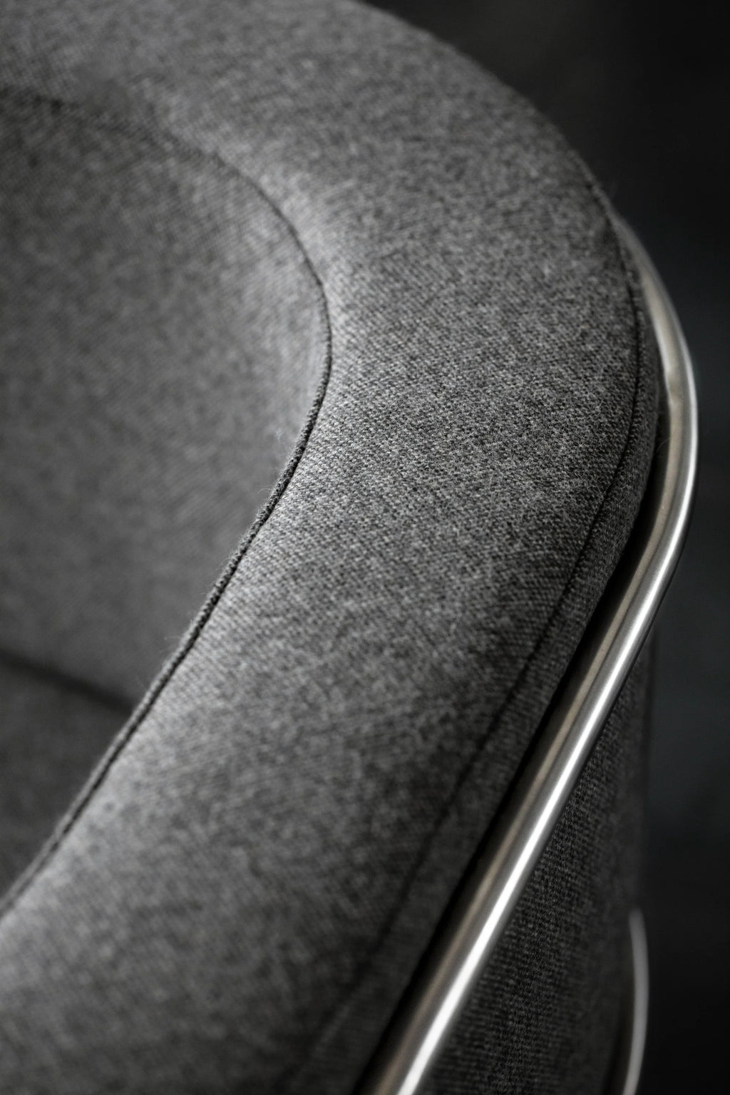 Kristina Dam Modernist Lounge Chair, a sleek contemporary lounge chair featuring a dark grey textured fabric upholstery with clean lines and a gently curved silhouette. Its minimalist design is accentuated by a contrasting brushed metal frame that elegantly traces the chair's back and armrest. Ideal as a stylish accent armchair for modern living rooms, Scandinavian-inspired spaces, or cozy reading nooks.