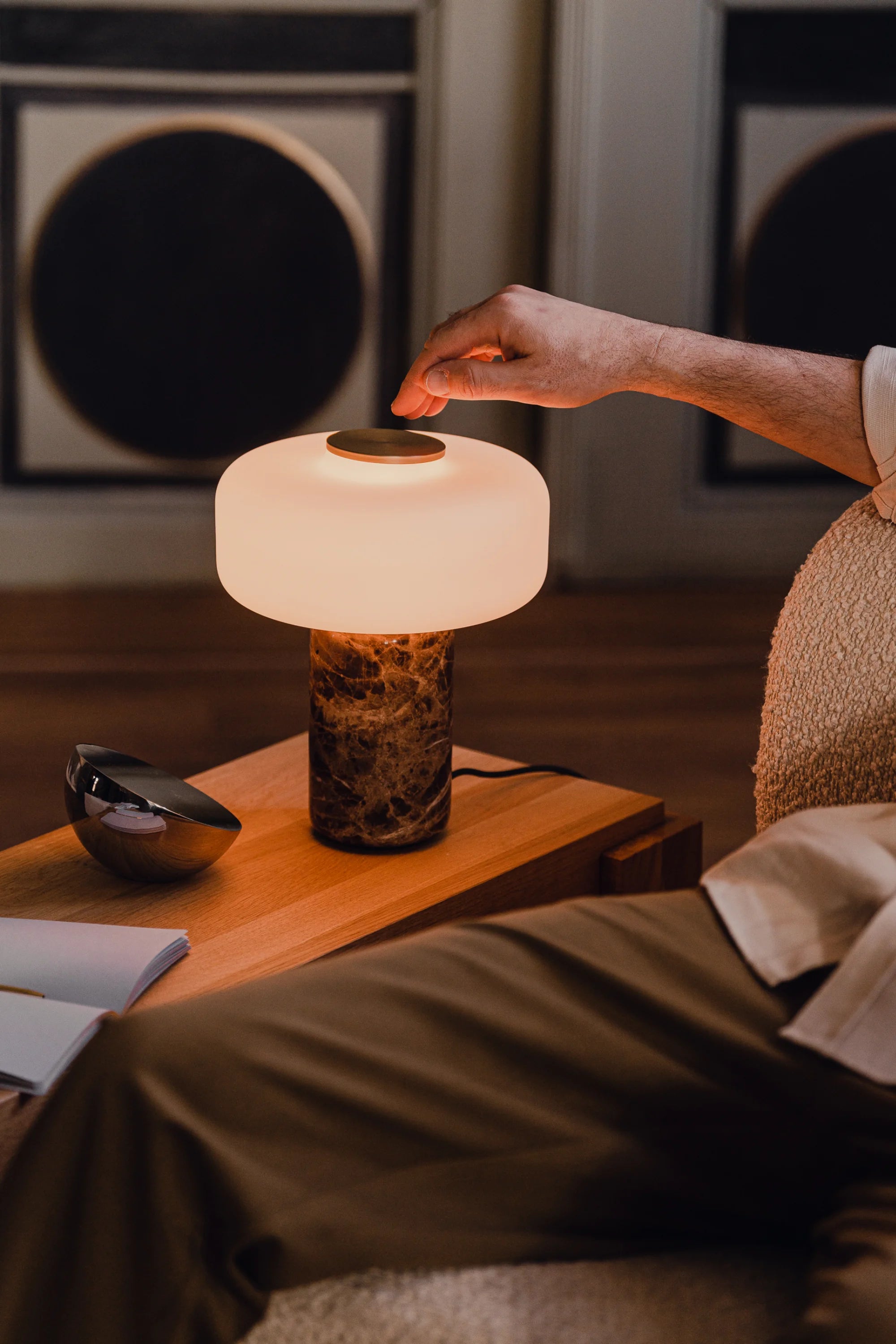 A person is adjusting the Tamo Table lamp on a wooden table with a cozy indoor setting.