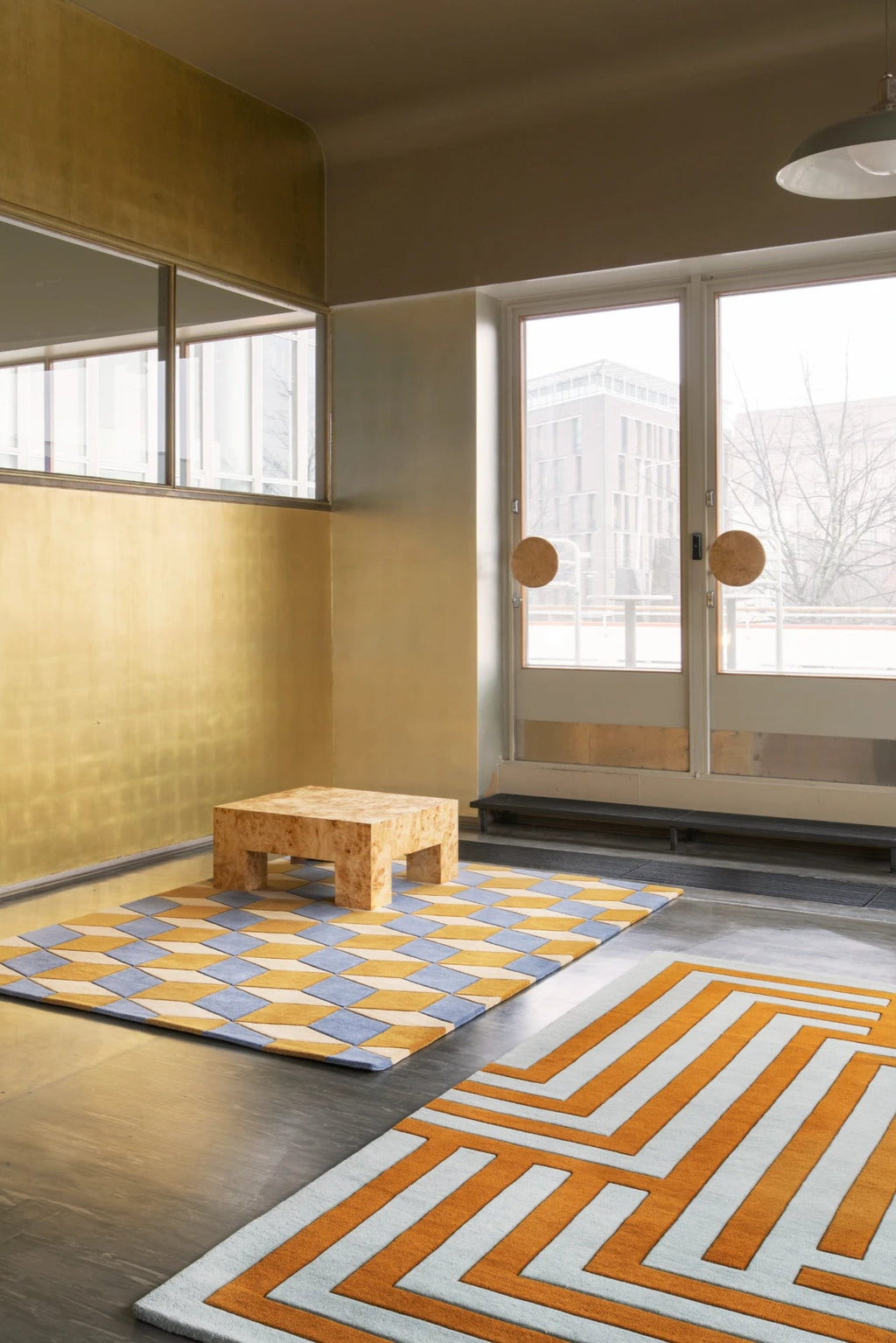 Layered Letters Wool Rug, featuring two distinct geometric pattern rugs in an interior setting. One rug displays a 3D cube effect with yellow, light blue, and cream diamonds, while the other featur...
